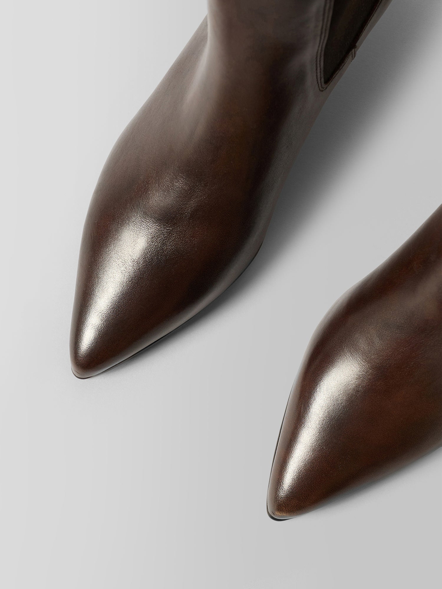 Close-up of a pair of pointed-toe ankle boots in dark brown leather, showing the smooth texture and polished finish on a light gray background.