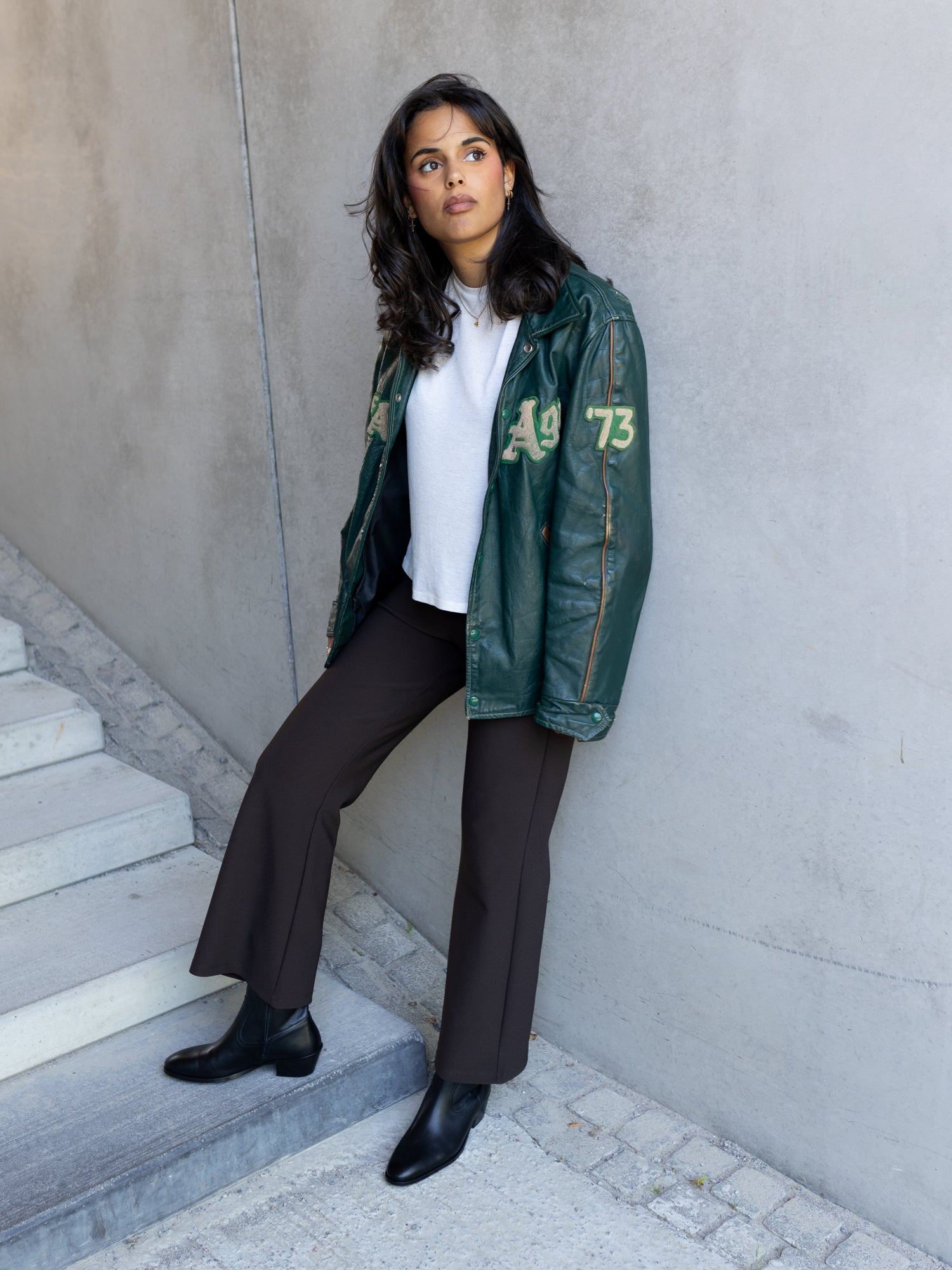A woman wearing a dark green leather jacket with yellow-green 'A9 73' lettering, a white top, dark brown wide-leg trousers, and black ankle boots, posing on concrete stairs against a gray wall.