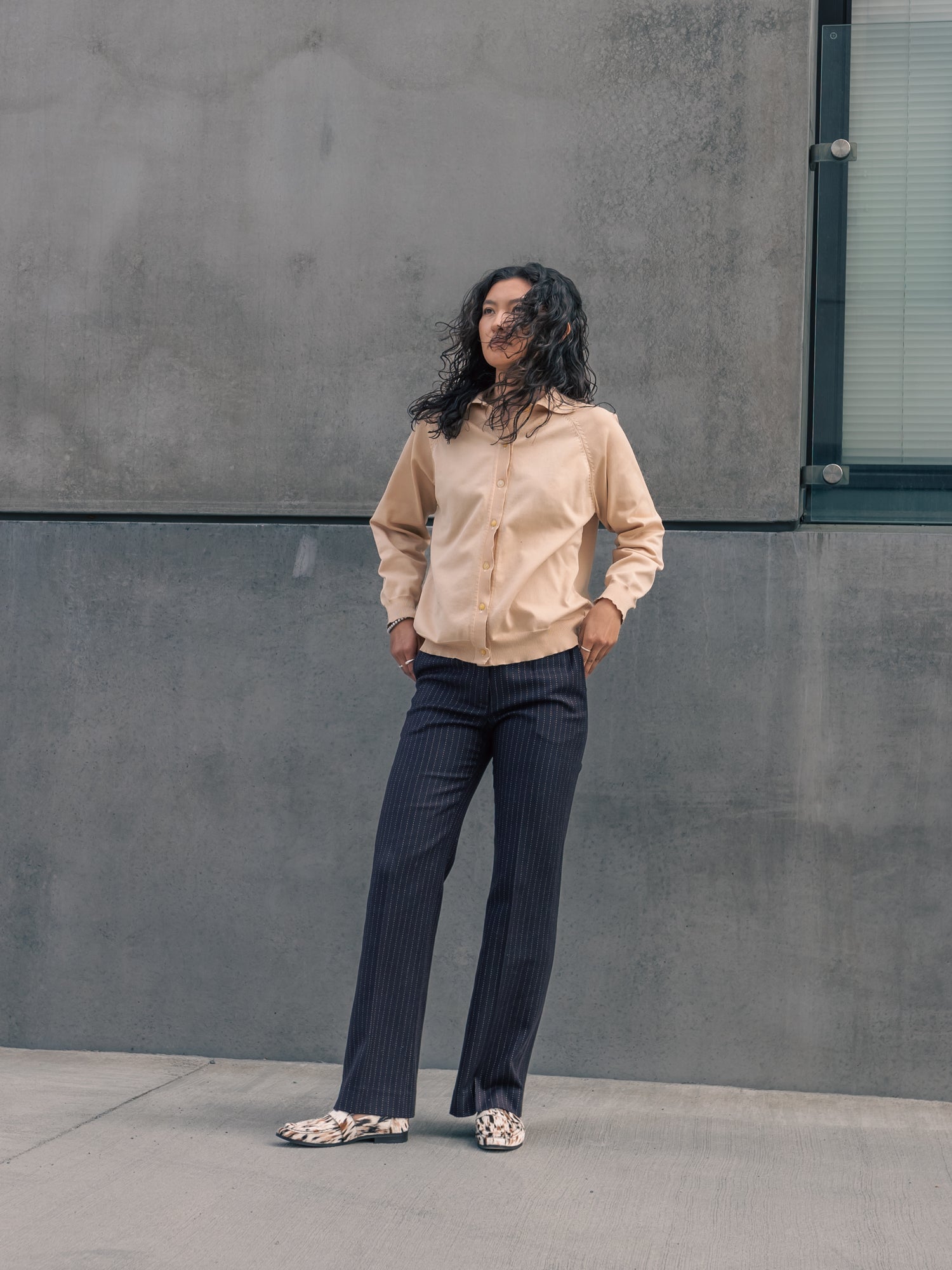 Person wearing a beige shirt and dark pants standing against a gray wall.